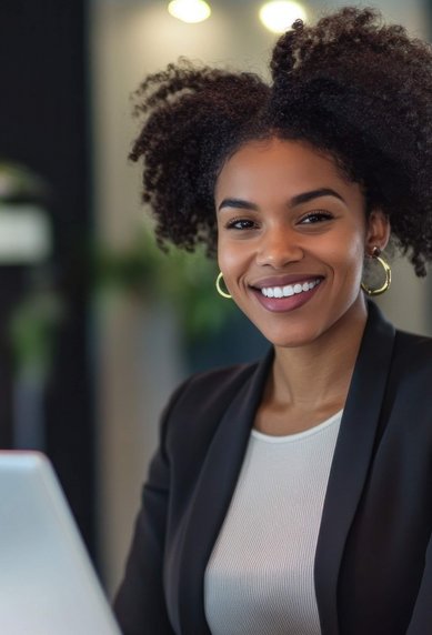 Eine Frau mit lockigem Haar sitzt an einem Empfangstresen und lächelt, während sie auf einen Laptop schaut. Pflanzen sind im Hintergrund sichtbar.