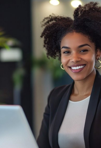A woman with curly hair sits at a reception desk, smiling as she looks at a laptop. Plants are visible in the background.