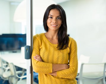Eine Frau mit langen, dunklen Haaren trägt einen gelben Pullover und steht an einer Glastür. Sie lächelt und hat die Arme vor der Brust verschränkt. Im Hintergrund sind Büromöbel und Bildschirme sichtbar.