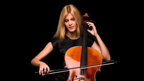 Eine junge Frau spielt Cello, während sie konzentriert auf die Saiten schaut. Sie hat lange, blonde Haare und trägt ein schwarzes Oberteil.