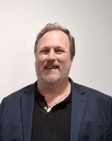 A man with medium-length gray hair and a beard is wearing a black polo shirt and a blue blazer. He is smiling warmly.