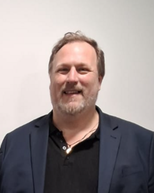 A man with medium-length gray hair and a beard is wearing a black polo shirt and a blue blazer. He is smiling warmly.