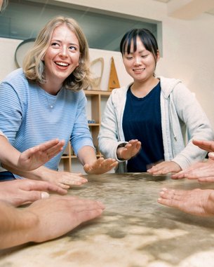 Sechs Personen stehen um einen Tisch und legen ihre Hände auf die Oberfläche. Einige lächeln, während sie sich auf eine gemeinsame Aktivität konzentrieren. Der Tisch hat eine glatte, helle Oberfläche.