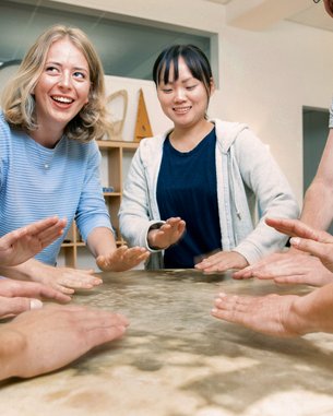Sechs Personen stehen um einen Tisch und legen ihre Hände auf die Oberfläche. Einige lächeln, während sie sich auf eine gemeinsame Aktivität konzentrieren. Der Tisch hat eine glatte, helle Oberfläche.