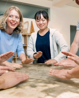 Sechs Personen stehen um einen Tisch und legen ihre Hände auf die Oberfläche. Einige lächeln, während sie sich auf eine gemeinsame Aktivität konzentrieren. Der Tisch hat eine glatte, helle Oberfläche.