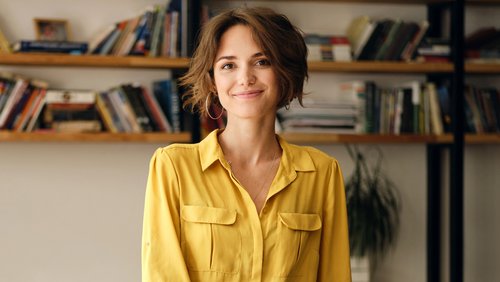 Eine Frau mit kurzen, lockigen Haaren trägt ein gelbes Hemd und lächelt. Im Hintergrund sind Bücherregale sichtbar.