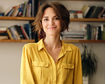 Eine Frau mit kurzen, lockigen Haaren trägt ein gelbes Hemd und lächelt. Im Hintergrund sind Bücherregale sichtbar.