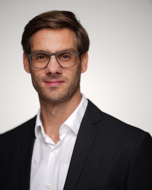 A man in glasses and a suit smiles at the camera. The background is blurred and bright, drawing attention to his face.
