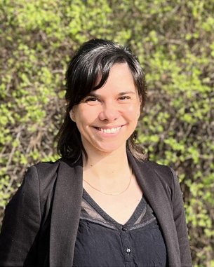 Eine Frau mit dunklen Haaren und einem schwarzen Blazer lächelt vor einem Hintergrund aus grünen Pflanzen.