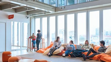 A group of five people is sitting on large orange bean bags in a modern room with big windows.