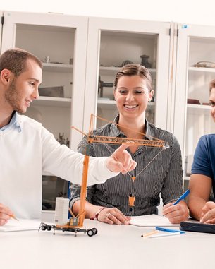 Drei Personen sitzen an einem Tisch und diskutieren. Eine Person zeigt auf ein Modell eines Krans, während die anderen aufmerksam zuhören. Auf dem Tisch liegen Schreibmaterialien und Notizen.