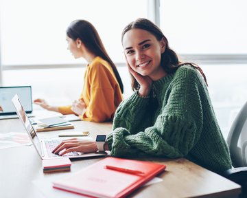 Eine lächelnde Frau mit einem grünen Pullover sitzt an einem Tisch mit einem Laptop, während eine andere Person im Hintergrund arbeitet.