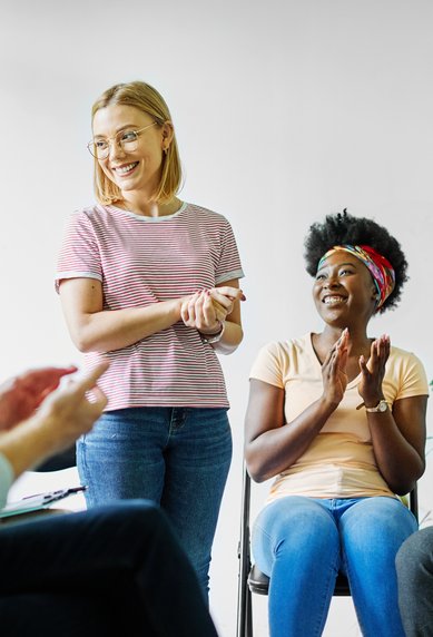 Eine Gruppe von Menschen sitzt in einem Raum. Eine Frau steht lächelnd im Mittelpunkt und wird von den anderen applaudiert. Pflanzen sind im Hintergrund sichtbar.
