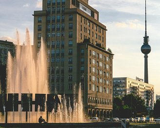 Ein Brunnen mit Wasserfontänen im Vordergrund, umgeben von modernen Gebäuden. Im Hintergrund ist der Fernsehturm von Berlin sichtbar, der sich gegen den Himmel abhebt.