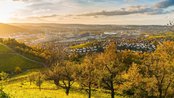 A picturesque view of a city in autumn, surrounded by colorful trees and gentle hills, as the sun sets and paints the sky in warm hues.