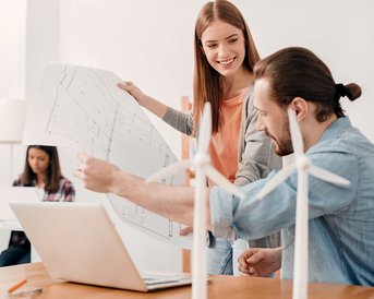 Zwei Personen besprechen Baupläne am Tisch, während eine dritte Person im Hintergrund an einem Laptop arbeitet. Auf dem Tisch stehen kleine Windturbinenmodelle.