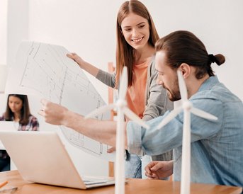 Two people are discussing construction plans at the table, while a third person in the background is working on a laptop. On the table, there are small models of wind turbines.