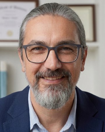 Man with glasses and a gray beard, smiling in a suit in front of a bookshelf with certificates in the background.
