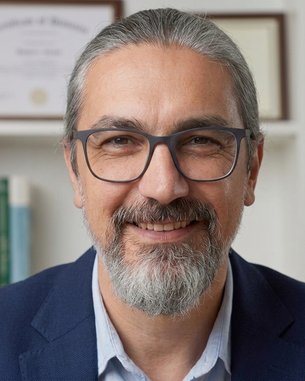 Man with glasses and a gray beard, smiling in a suit in front of a bookshelf with certificates in the background.