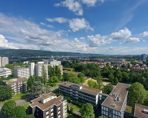 Blick auf eine Stadtlandschaft mit modernen Wohngebäuden, viel Grün und einem Hügel im Hintergrund unter einem klaren Himmel mit einigen Wolken.