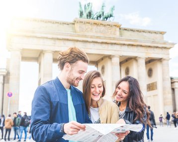 Drei Personen stehen vor dem Brandenburger Tor und betrachten eine Karte, während sie lächeln und miteinander sprechen.
