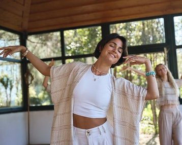Two women are dancing in a bright room with large windows. One woman is wearing a white top and a light jacket, while the other is dressed in a similar outfit.
