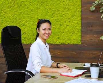 A young woman is sitting at a desk in a modern office, smiling and working on a laptop. On the table, there’s a notebook, a smartphone, and a coffee cup. In the background, there’s a green wall with moss.