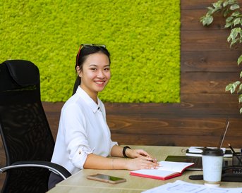 A young woman is sitting at a desk in a modern office, smiling and working on a laptop. On the table, there’s a notebook, a smartphone, and a coffee cup. In the background, there’s a green wall with moss.