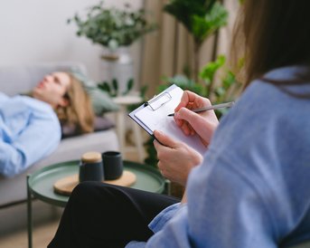 Eine Person sitzt mit einem Notizblock und schreibt, während eine andere Person entspannt auf einer Couch liegt. Im Hintergrund sind Pflanzen und eine Tasse zu sehen.