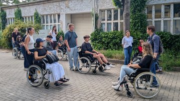 A group of young people in wheelchairs is standing on a sidewalk in front of a building, chatting and interacting with each other.