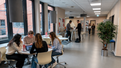 Studierende arbeiten an Tischen in einem modernen Raum, während andere sich im Hintergrund unterhalten oder stehen. Pflanzen sind ebenfalls sichtbar.