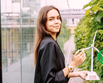 Eine junge Frau hält ein Modell einer Windkraftanlage in der Hand und lächelt, während sie vor einer grünen Wand steht.