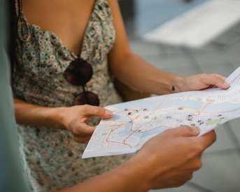 Eine Person hält eine gefaltete Karte in den Händen, während eine andere Person daneben steht und auf die Karte schaut. Beide scheinen sich in einer städtischen Umgebung zu befinden.