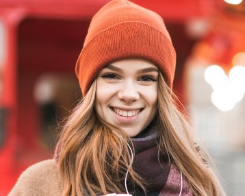 Junge Frau mit braunen Haaren trägt einen orangefarbenen Hut und hält einen grünen Kaffeebecher in der Hand, lächelt vor einem roten Hintergrund.
