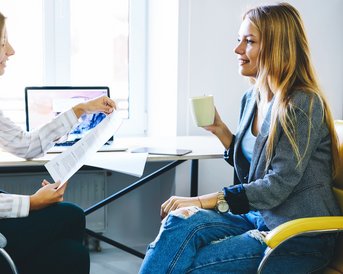 Zwei Frauen sitzen in einem modernen Büro. Eine zeigt auf ein Dokument, während die andere eine Tasse hält und aufmerksam zuhört.