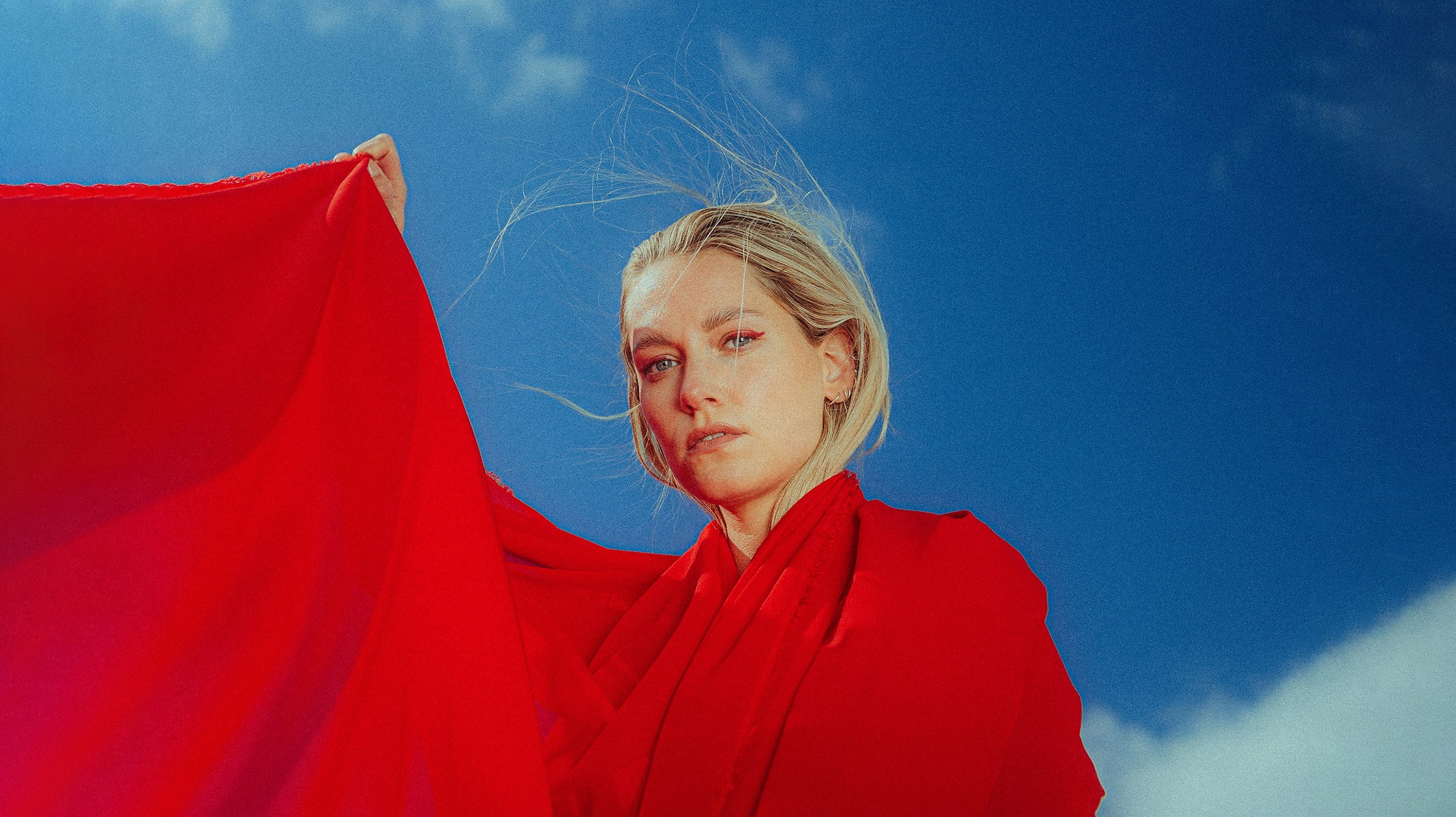 A person with blonde hair is wearing a red fabric and standing against a clear blue sky. The fabric flutters in the wind as the person looks directly at the camera.