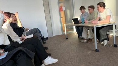 Three people are sitting at a table, while a group of listeners is seated across from them. A laptop is placed on the table.