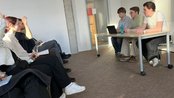 Drei Personen sitzen an einem Tisch, während eine Gruppe von Zuhörern ihnen gegenüber sitzt. Ein Laptop ist auf dem Tisch platziert.