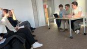 Three people are sitting at a table, while a group of listeners is seated across from them. A laptop is placed on the table.