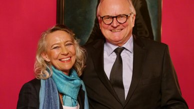Ein Mann und eine Frau stehen lächelnd vor einer pinken Wand, beide in formeller Kleidung. Der Mann trägt eine Brille und einen Anzug.