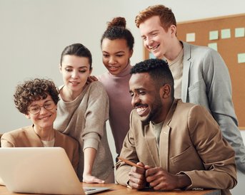 Fünf Personen stehen um einen Laptop und schauen gemeinsam auf den Bildschirm. Sie scheinen an einem Projekt oder einer Präsentation interessiert zu sein. Alle tragen legere, moderne Kleidung.