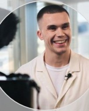 Ein junger Mann mit kurzem, glattem Haar und einem breiten Lächeln trägt ein helles Hemd. Er steht vor einer Kamera in einem hellen Raum.