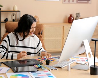 A person with long, dark hair is sitting at a desk, working on a computer, surrounded by color patterns and art supplies.