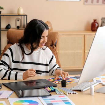 Eine Person mit langen, dunklen Haaren sitzt an einem Schreibtisch, arbeitet an einem Computer und umgeben von Farbmustern und Zeichenmaterialien.