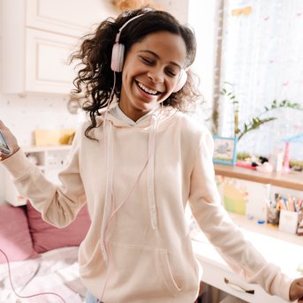 A girl with headphones smiles while holding a smartphone in her hand. The room is bright and cheerfully decorated.