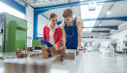 A woman in a red blazer and a man in blue work clothes are examining parts together on a table in a workshop.