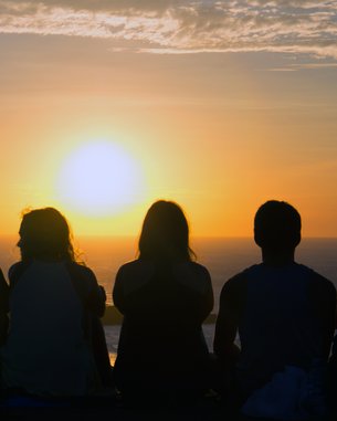 Six people are sitting side by side, watching the sunset on the horizon as the sky glows in warm colors.