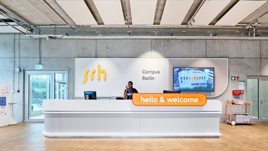 Modern reception desk at the SRH Campus Berlin with an employee welcoming visitors. A screen displays information about the campus.