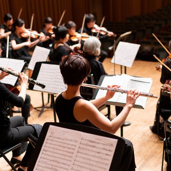Ein Orchester mit Musikerinnen und Musikern, die verschiedene Instrumente spielen, darunter Flöten und Violinen, während ein Dirigent dirigiert.