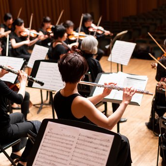 Ein Orchester mit Musikerinnen und Musikern, die verschiedene Instrumente spielen, darunter Flöten und Violinen, während ein Dirigent dirigiert.